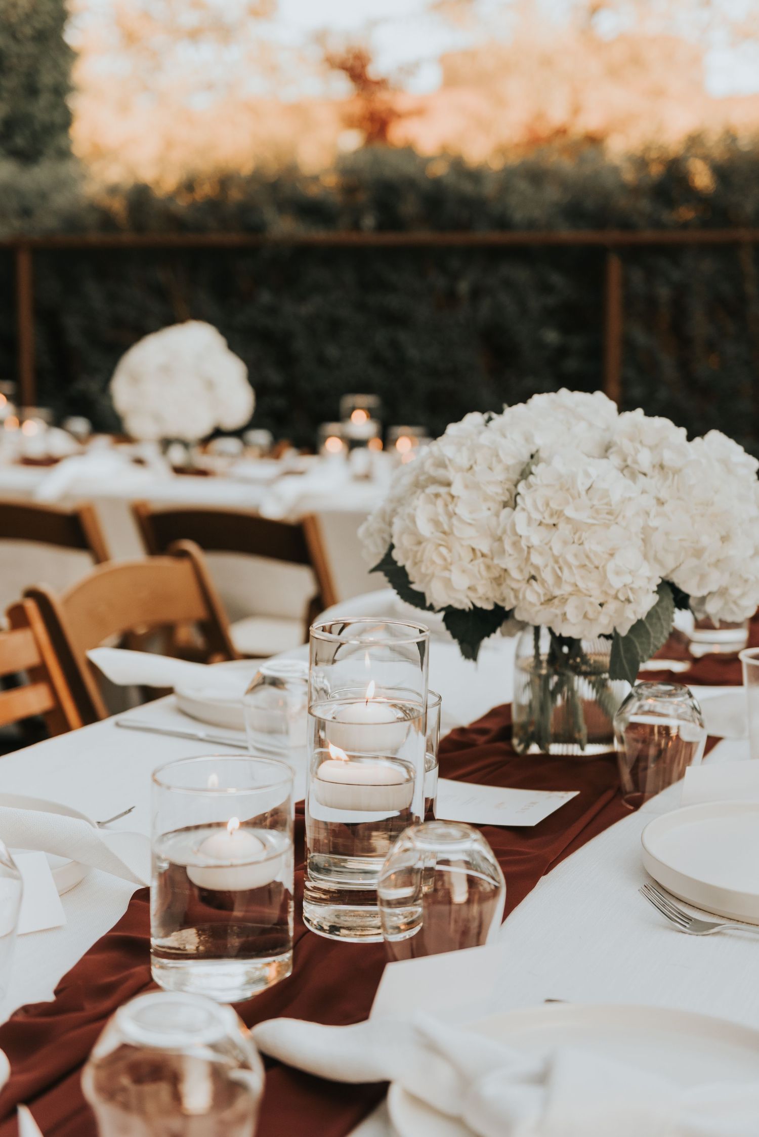 Rustic outdoor wedding reception table setup with white hydrangeas, burgundy runners, and simple glassware.