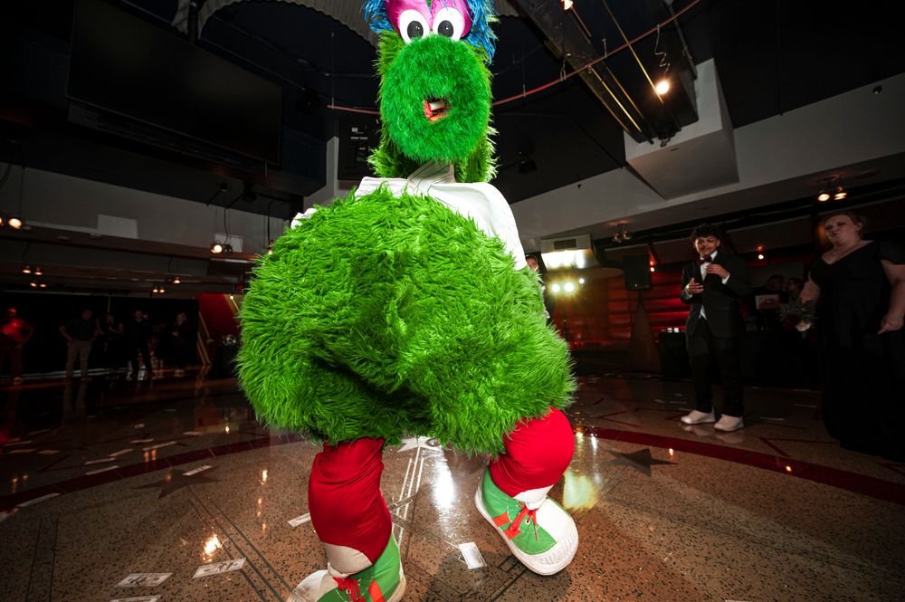 Green Phillie Phanatic mascot performs with red boxing gloves in a dark stadium setting.