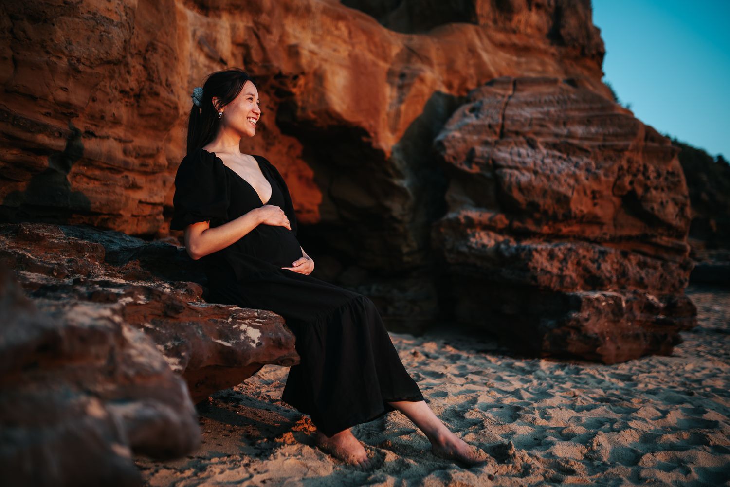 A pregnant person in a black dress sits on rock formations against a vibrant blue sky at sunset taken by Melbourne maternity photographer.