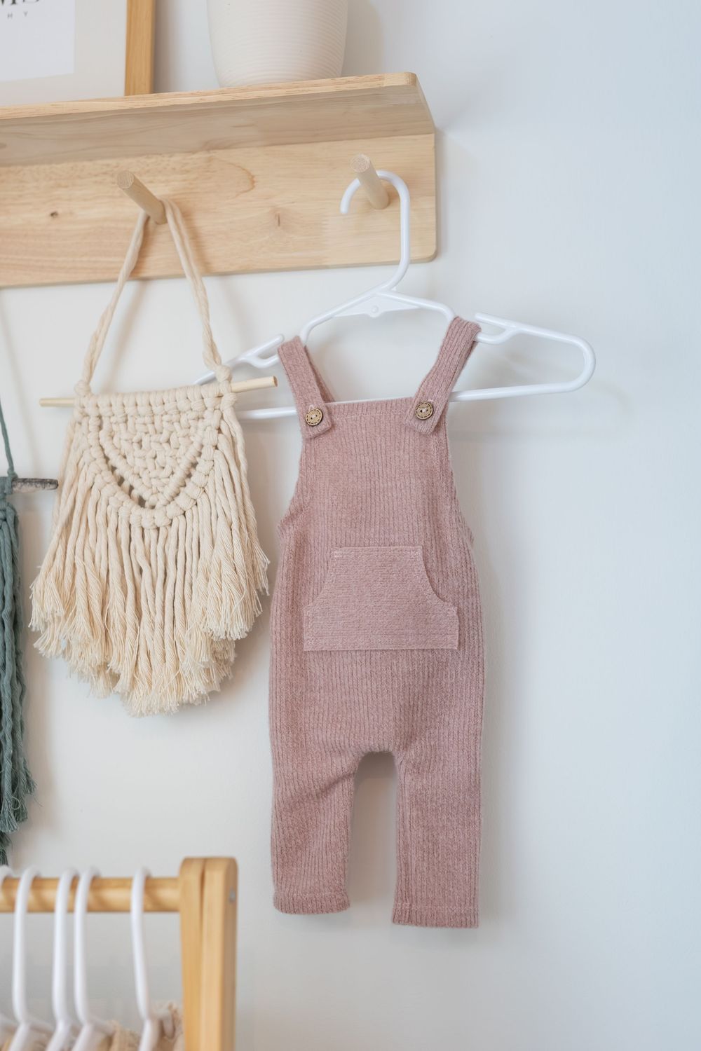 Boho baby clothing display with pink knit overalls and cream macrame wall hangings on wooden hooks.