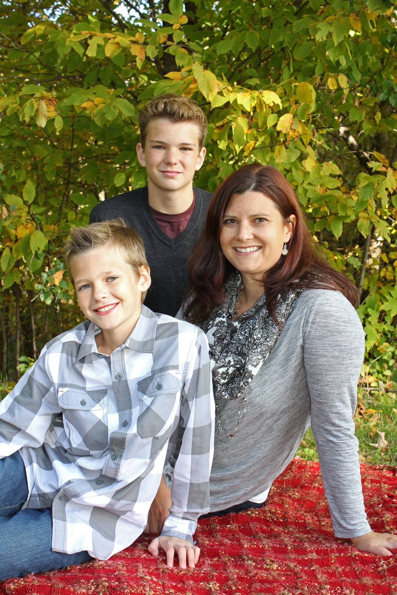 A woman and two boys smile together on a red blanket outdoors amid autumn foliage.