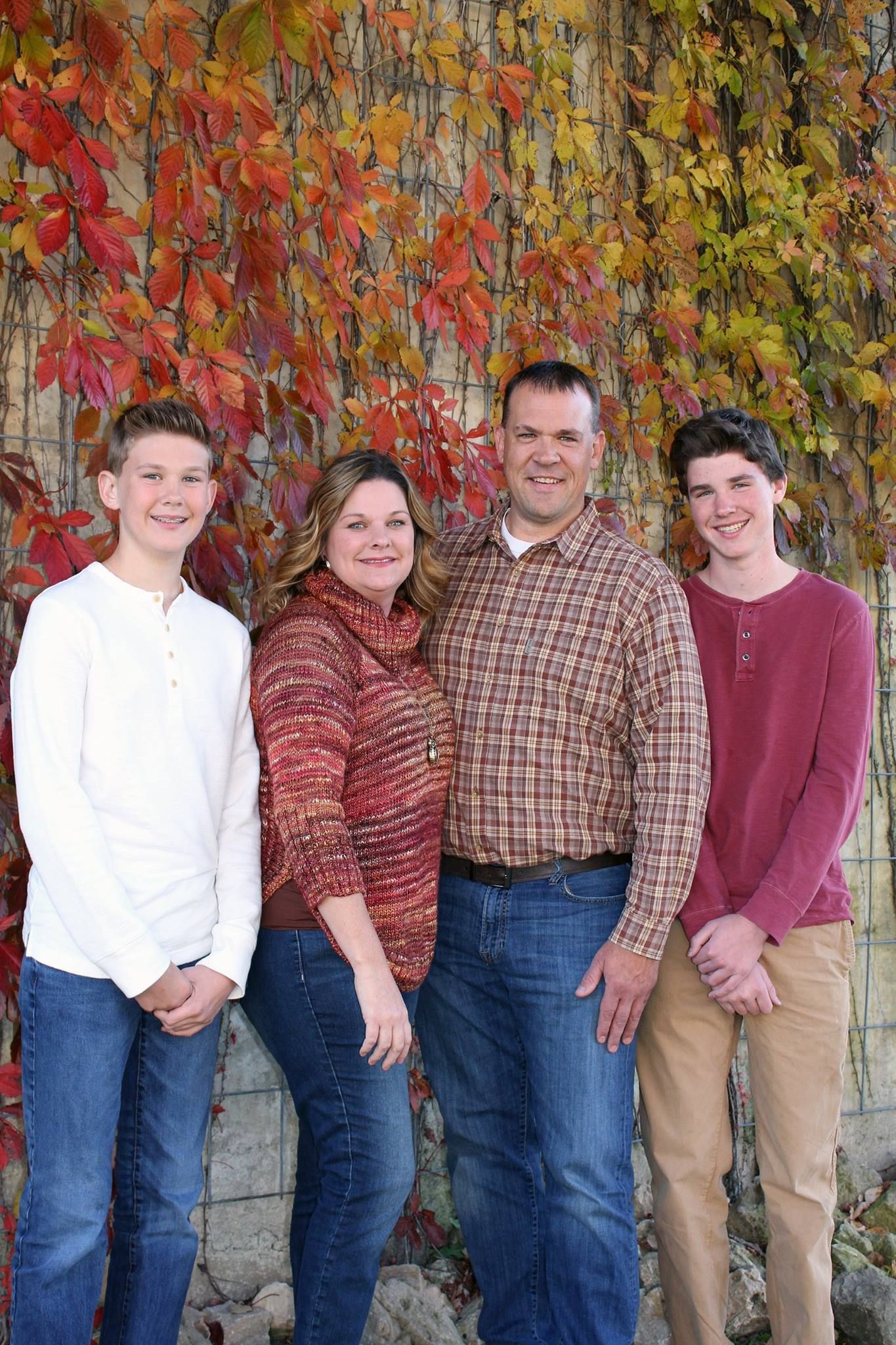 Family of four posing outdoors in fall, with colorful autumn foliage backdrop, wearing casual warm-toned clothing.