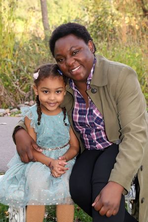A smiling woman and young girl in a teal dress pose together on a bench outdoors in a lush green park setting.