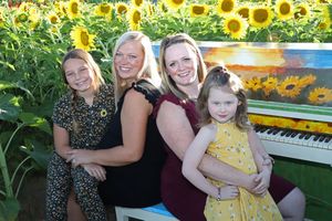 Two women and two girls pose happily in a sunflower field beside a painted piano on a sunny day.