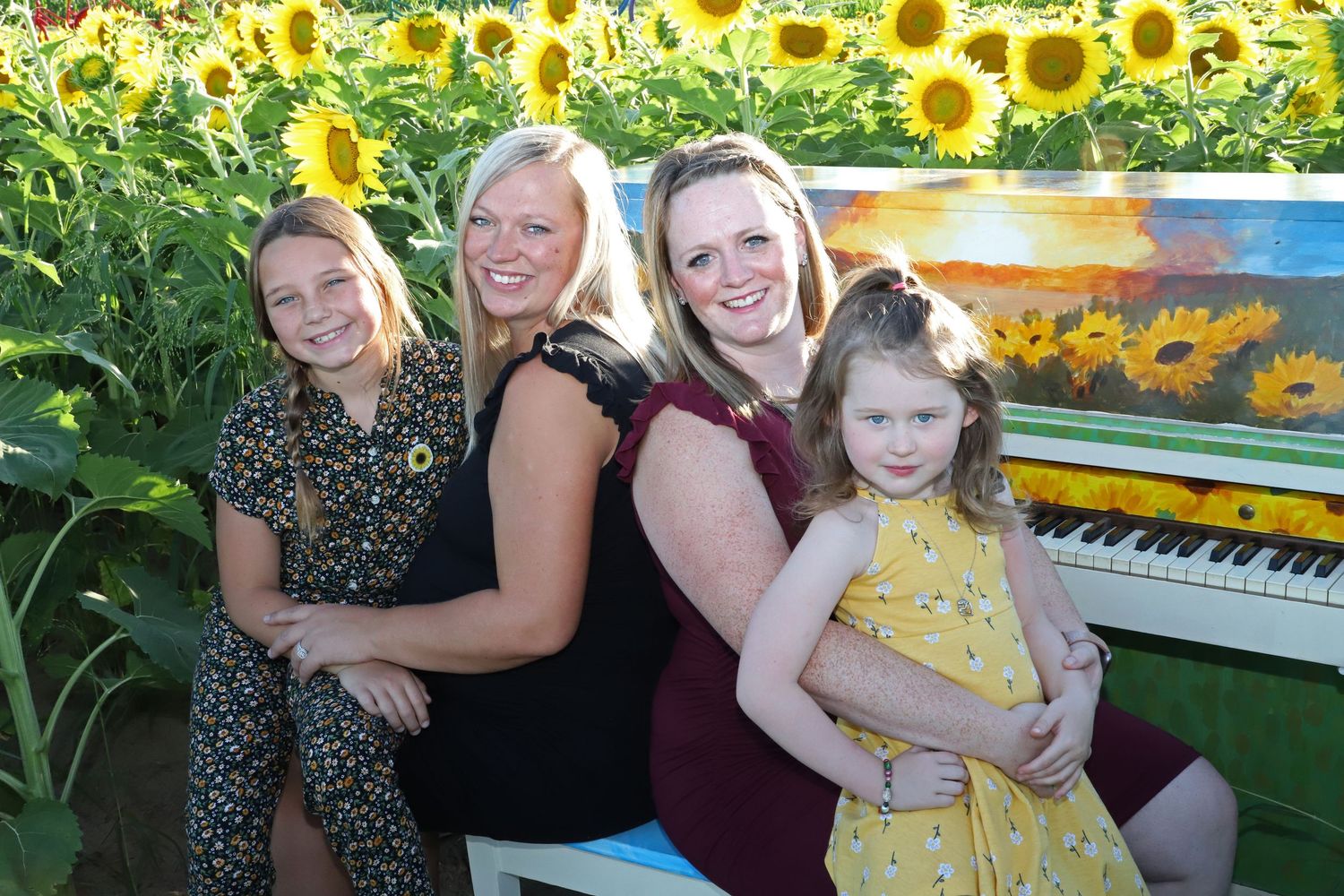 Two women and two girls pose happily in a sunflower field beside a painted piano on a sunny day.