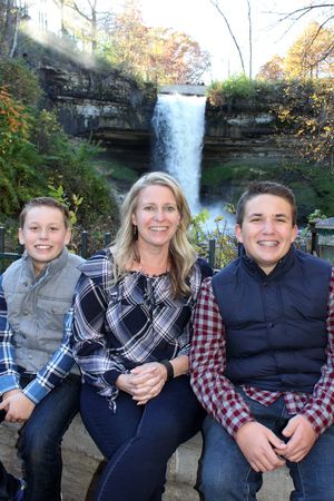 A woman and two boys smile together in front of a scenic waterfall surrounded by autumn foliage.