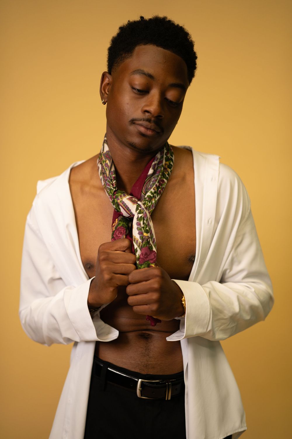 Black male model in open white shirt adjusting colorful neck scarf on yellow background.