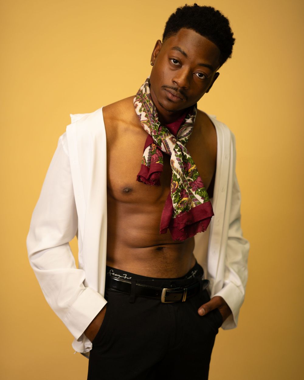 Young Black man in white blazer with floral scarf posing confidently on yellow background.