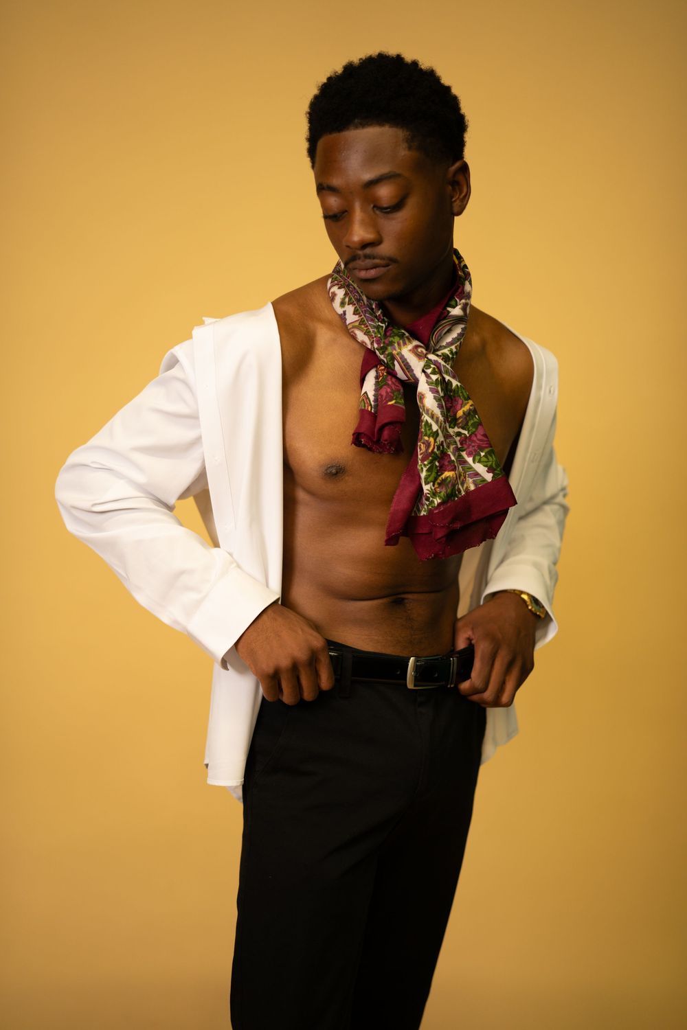 Black male model in open white blazer with colorful scarf and black pants on yellow background.