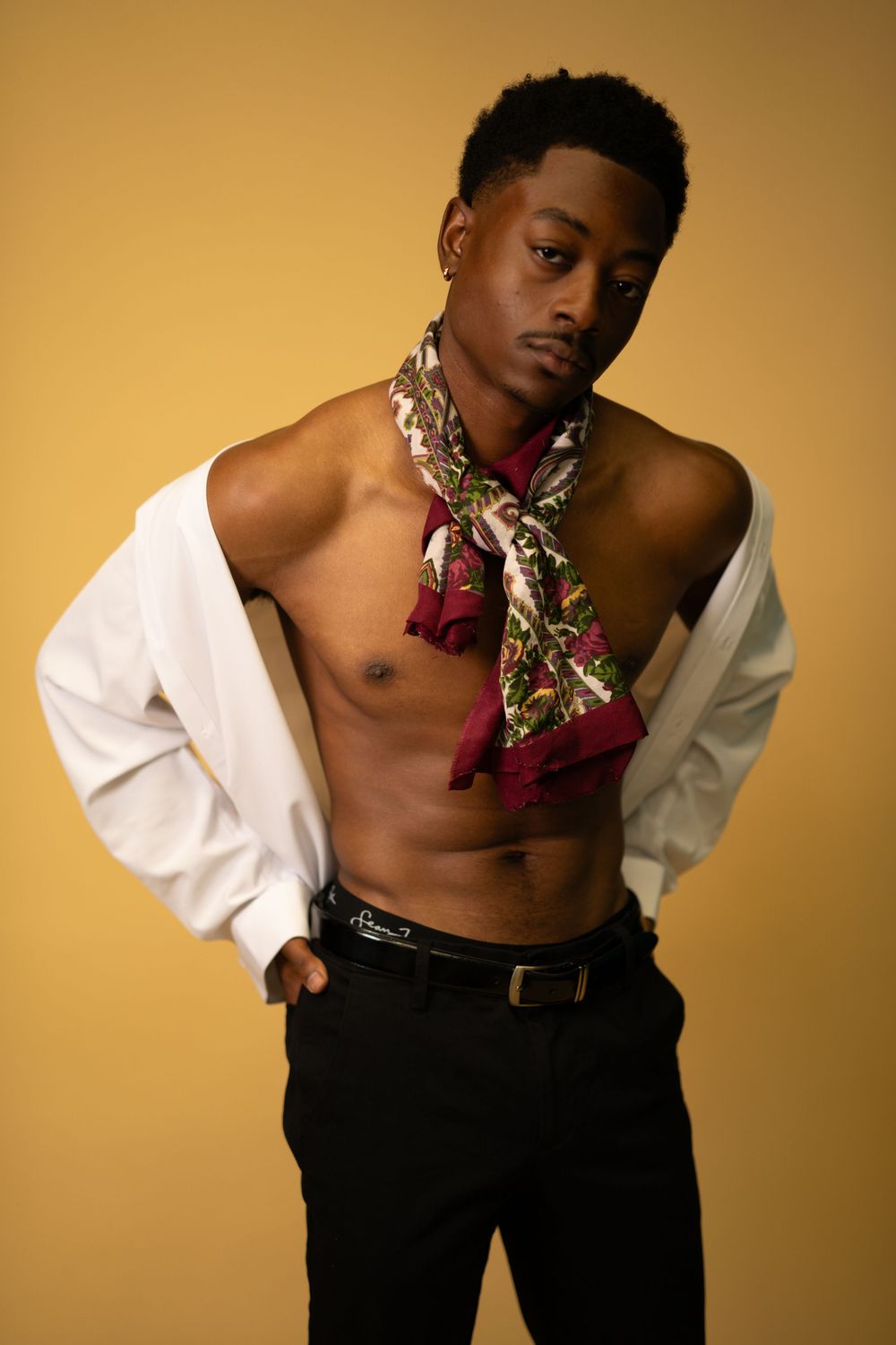 Black male model in white open shirt with colorful scarf posing on yellow background.