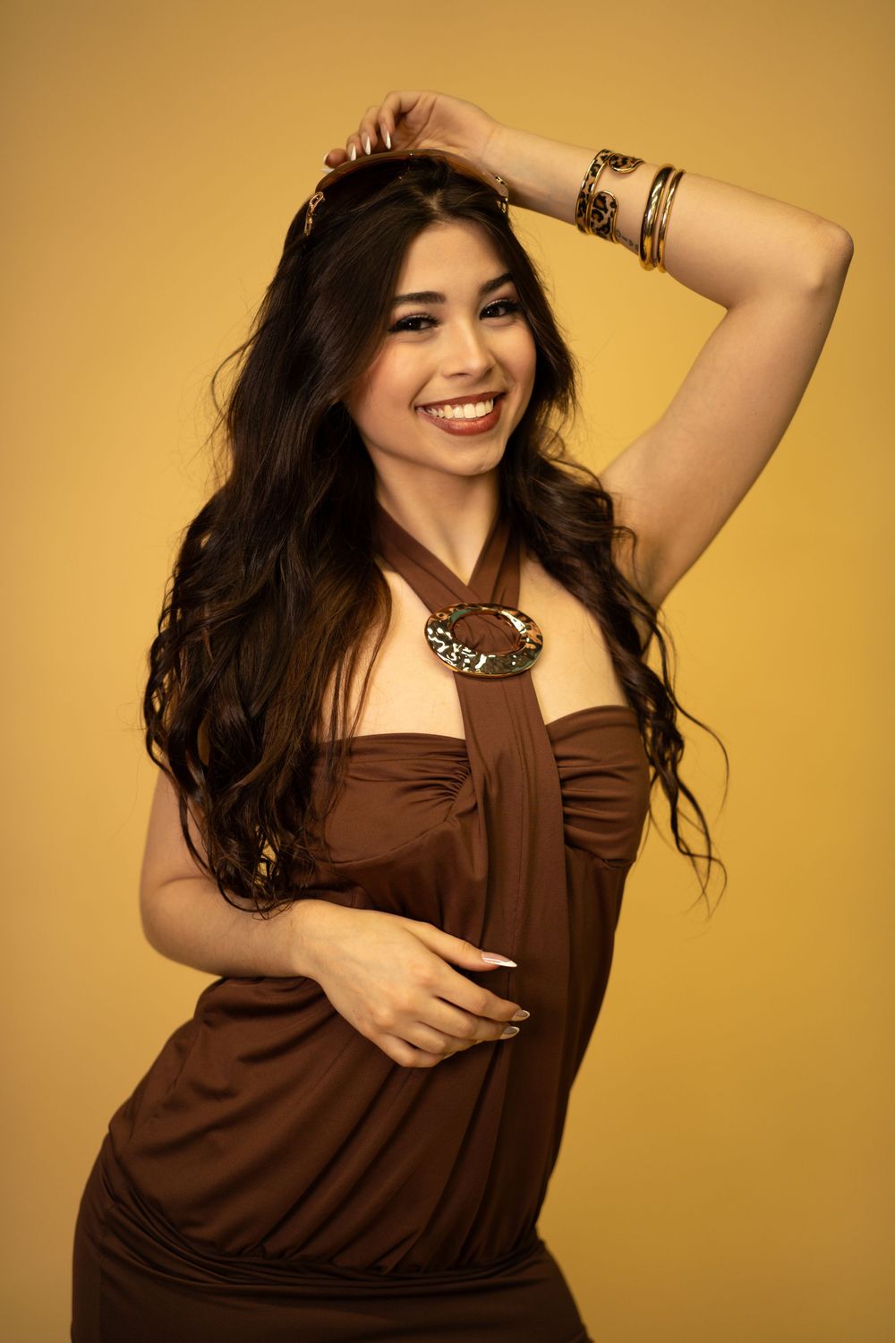 Smiling woman in brown halter dress with gold bracelets posing on yellow background.