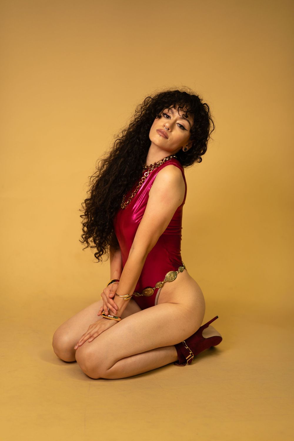 A woman with long curly hair poses in a red bodysuit and heels, kneeling on a yellow background.