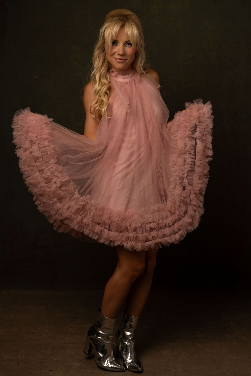 Blonde woman twirling in a pink ruffled tulle mini dress with silver ankle boots against a dark background.
