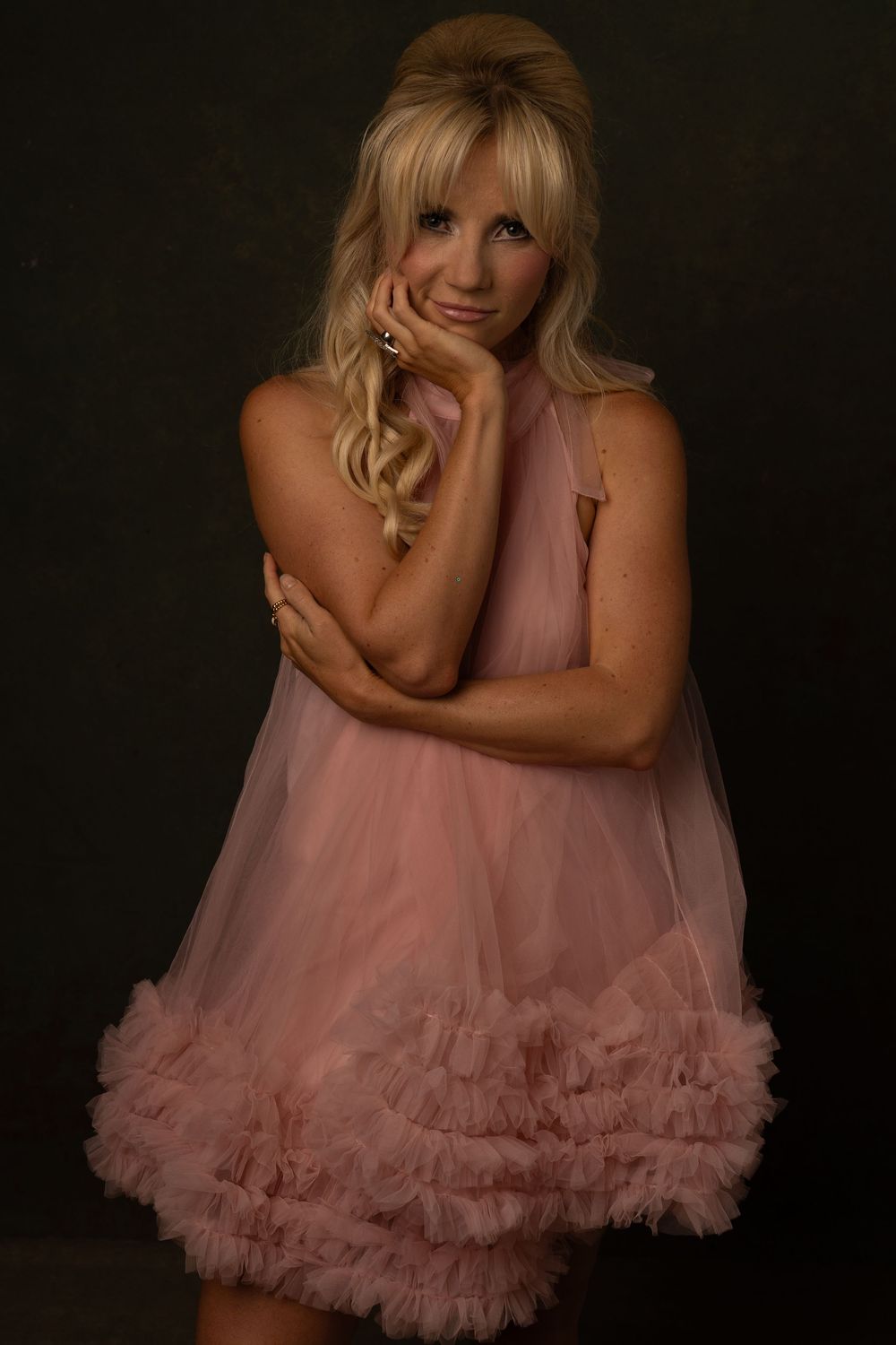 Blonde woman in a pink ruffled tulle dress poses thoughtfully with hand to cheek in studio.