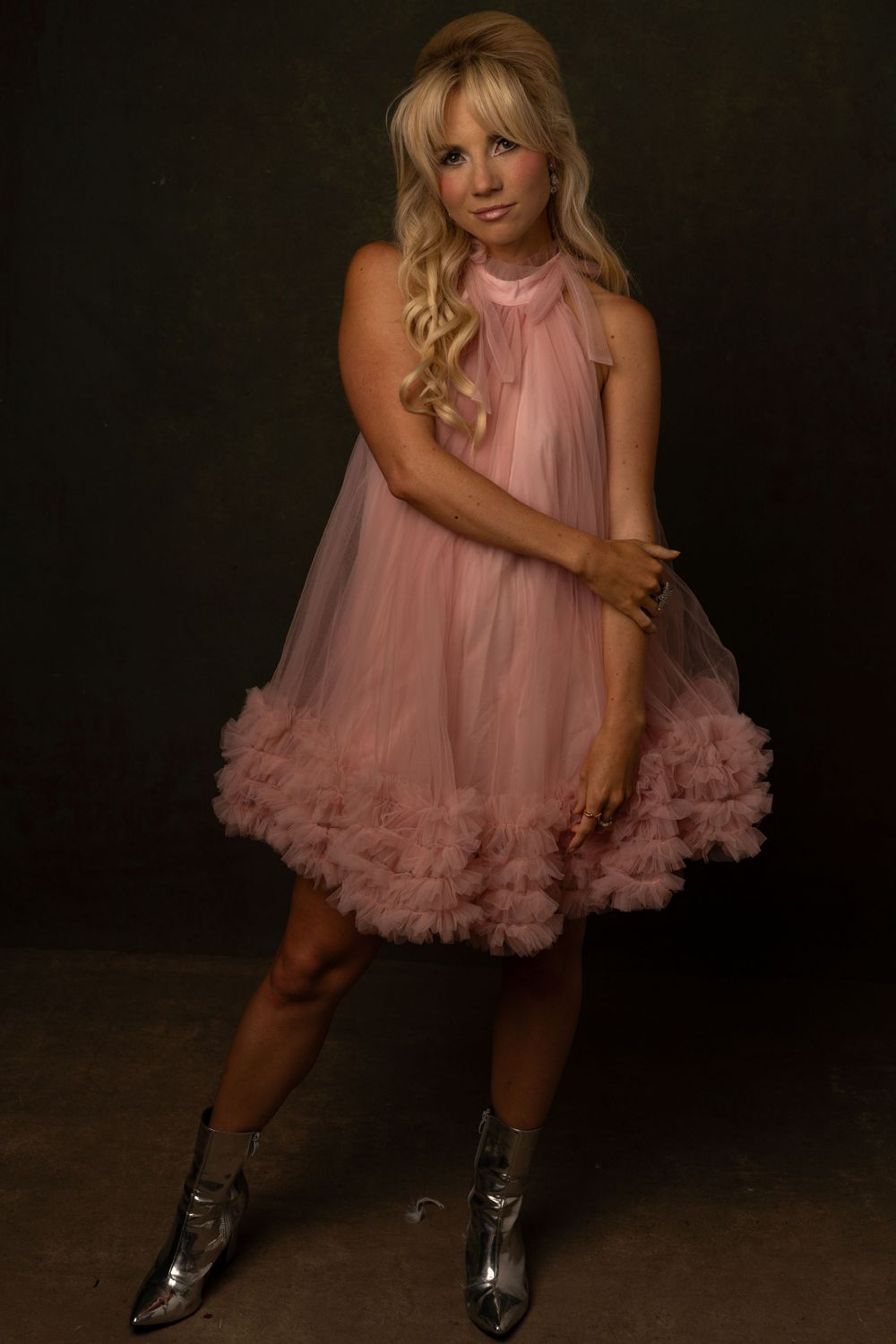 Blonde woman in pink ruffled tulle dress and silver boots poses dynamically in a dark studio.