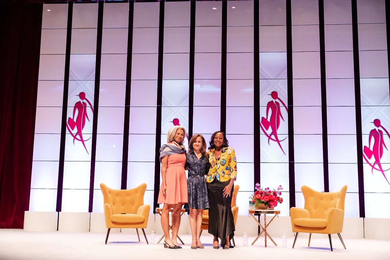 A minimalist stage design features orange chairs and vertical bars against a white backdrop with performers in silhouette.
