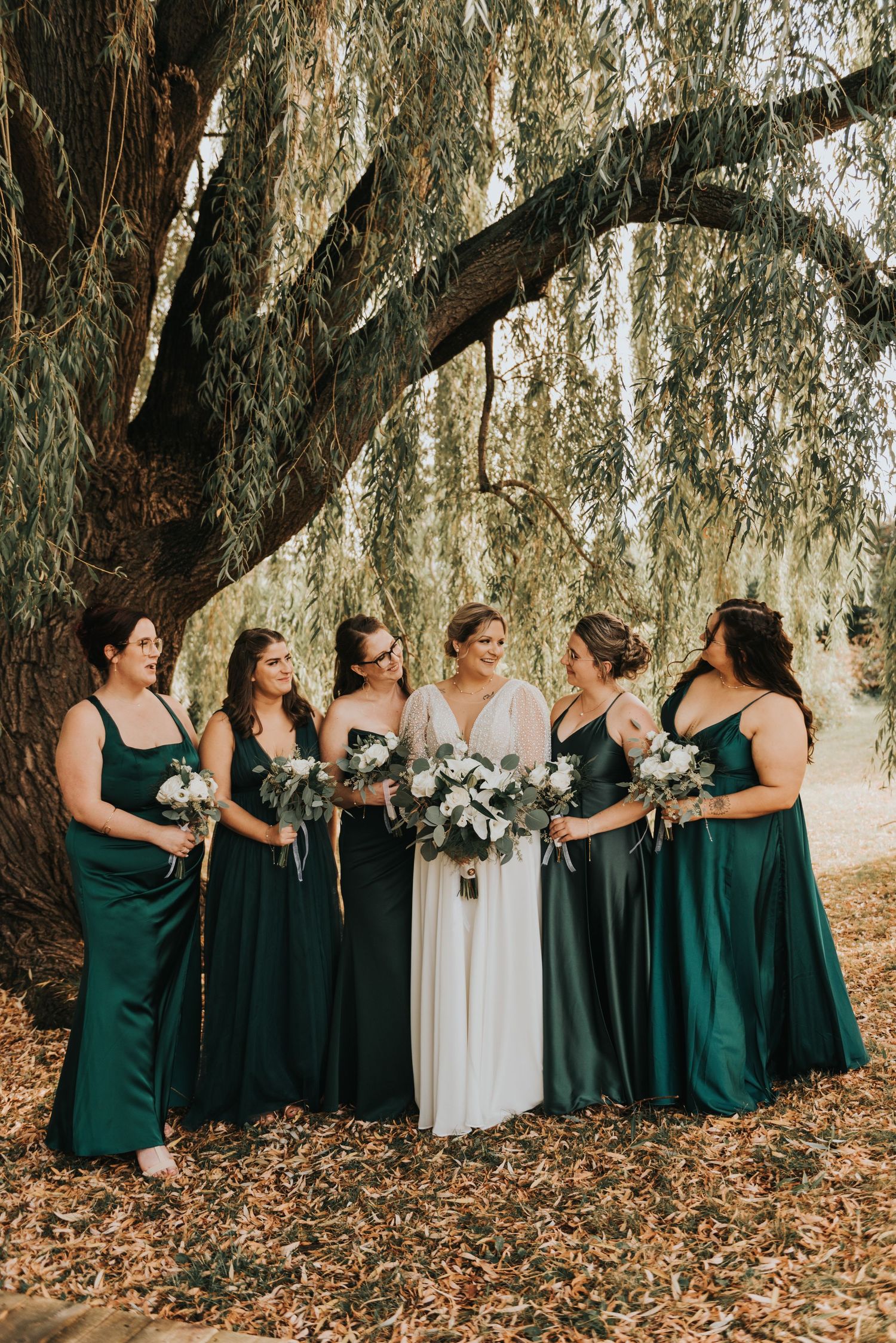 Wedding party in emerald green dresses stands together under willow trees on autumn day with fallen leaves.
