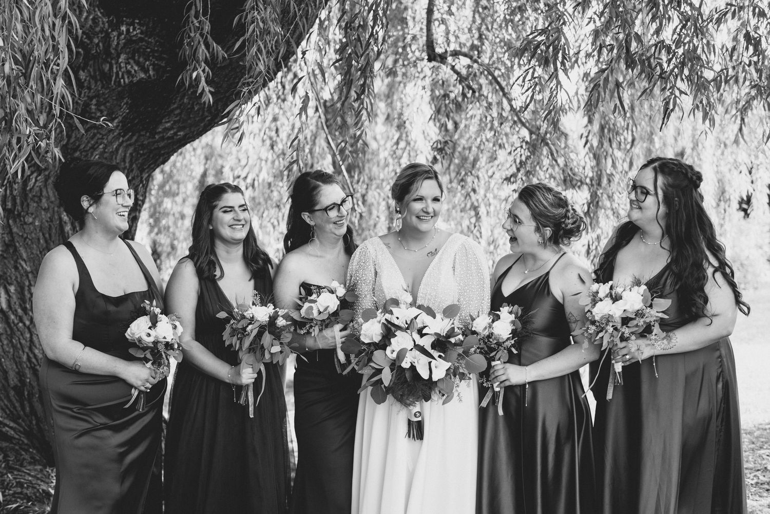 Wedding party stands in a row outdoors wearing black dresses and holding white floral bouquets against a natural backdrop.