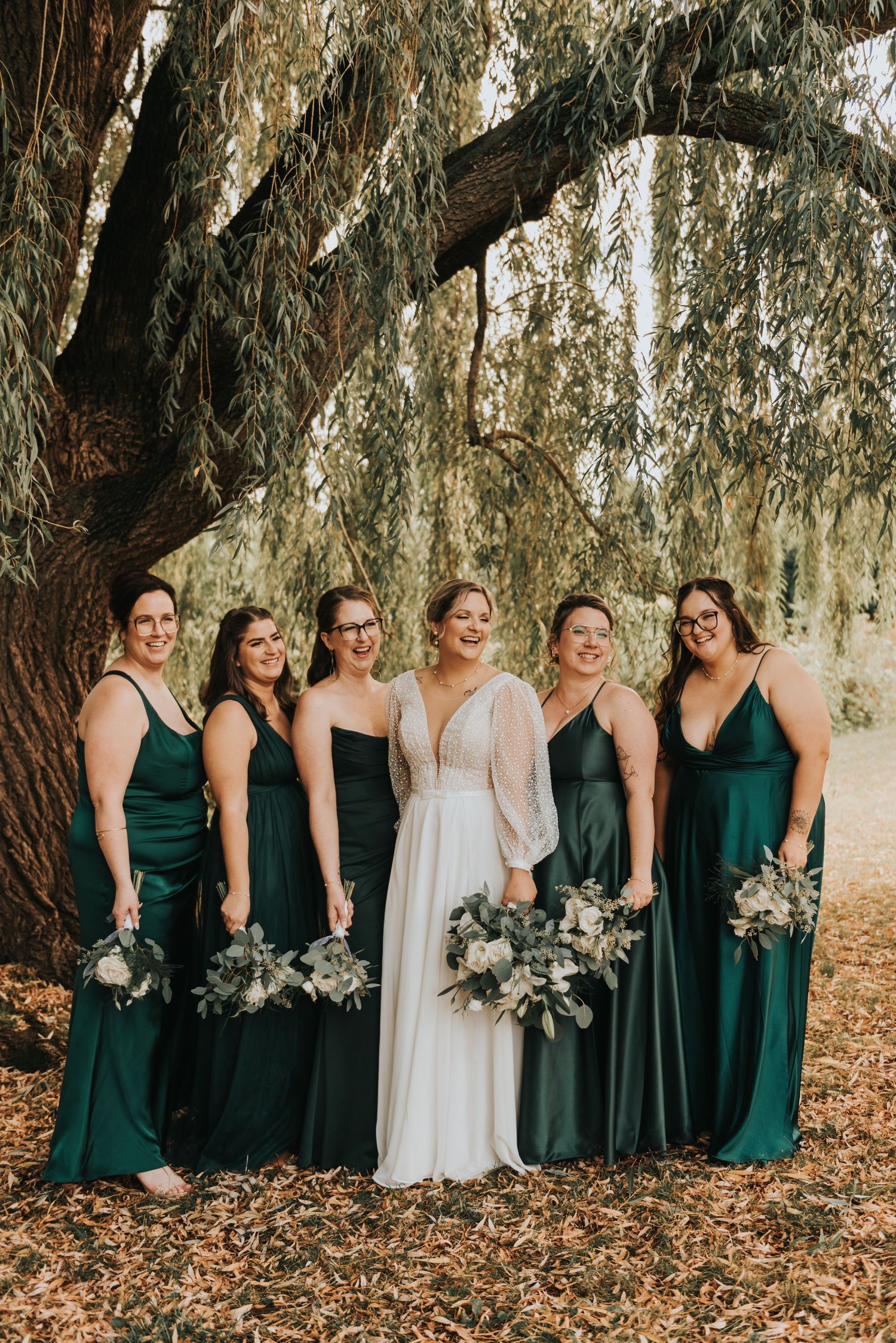 Wedding party in emerald green dresses poses under a weeping willow tree with autumn leaves on the ground.