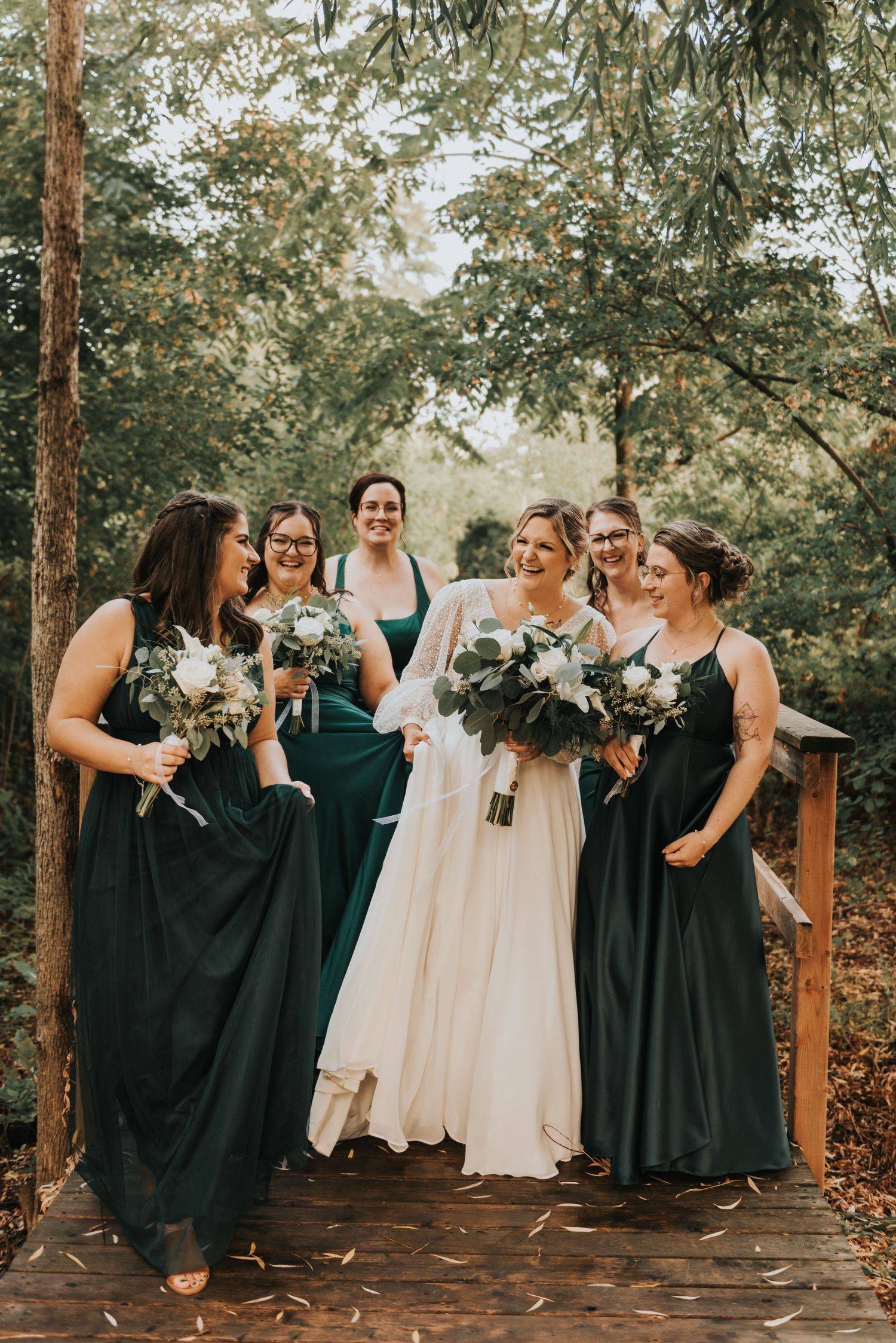 A bridal party in dark emerald dresses and white gown pose together in a wooded outdoor setting.