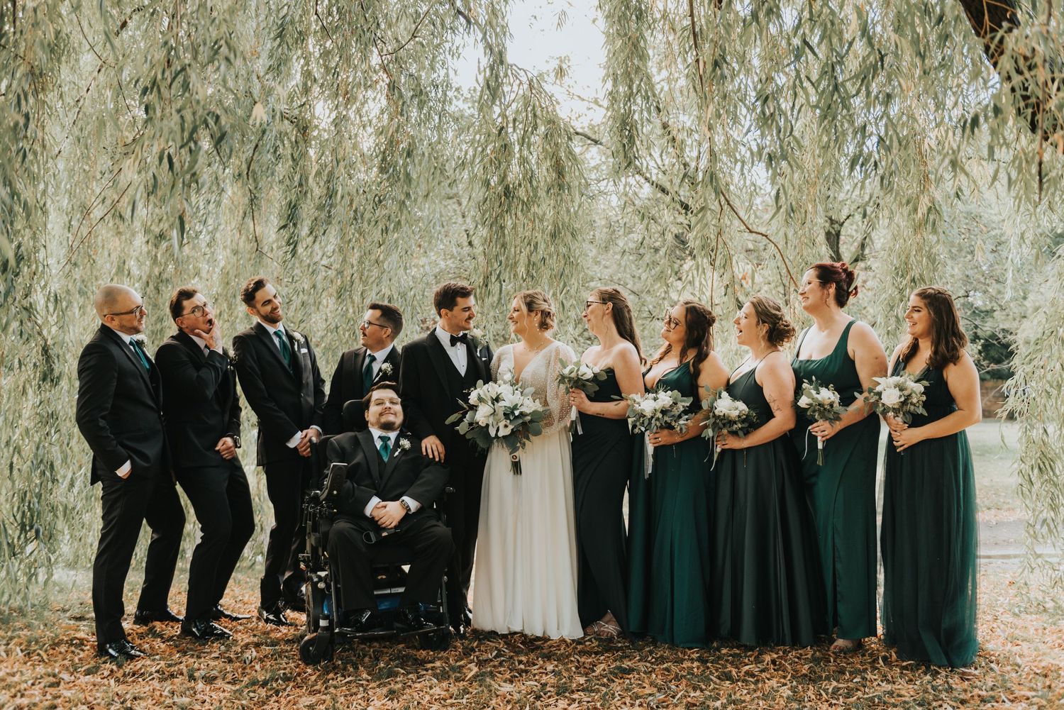 Wedding party poses together outdoors in a natural setting with trees and greenery.