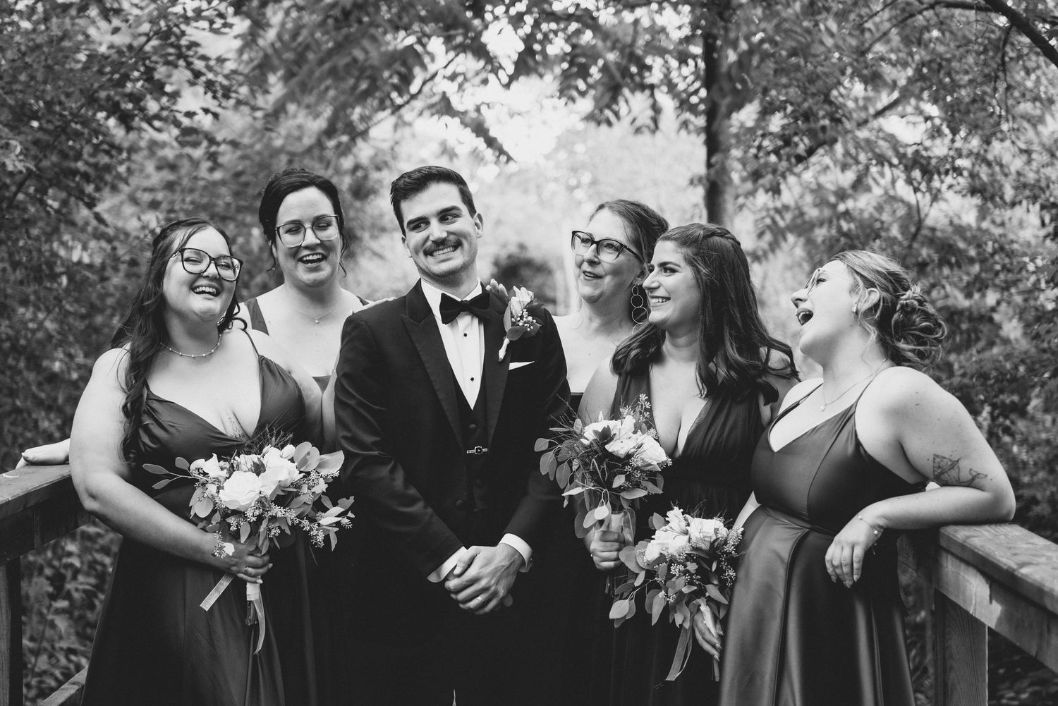 A joyful wedding party poses together outdoors in a garden setting with trees and natural greenery in the background.