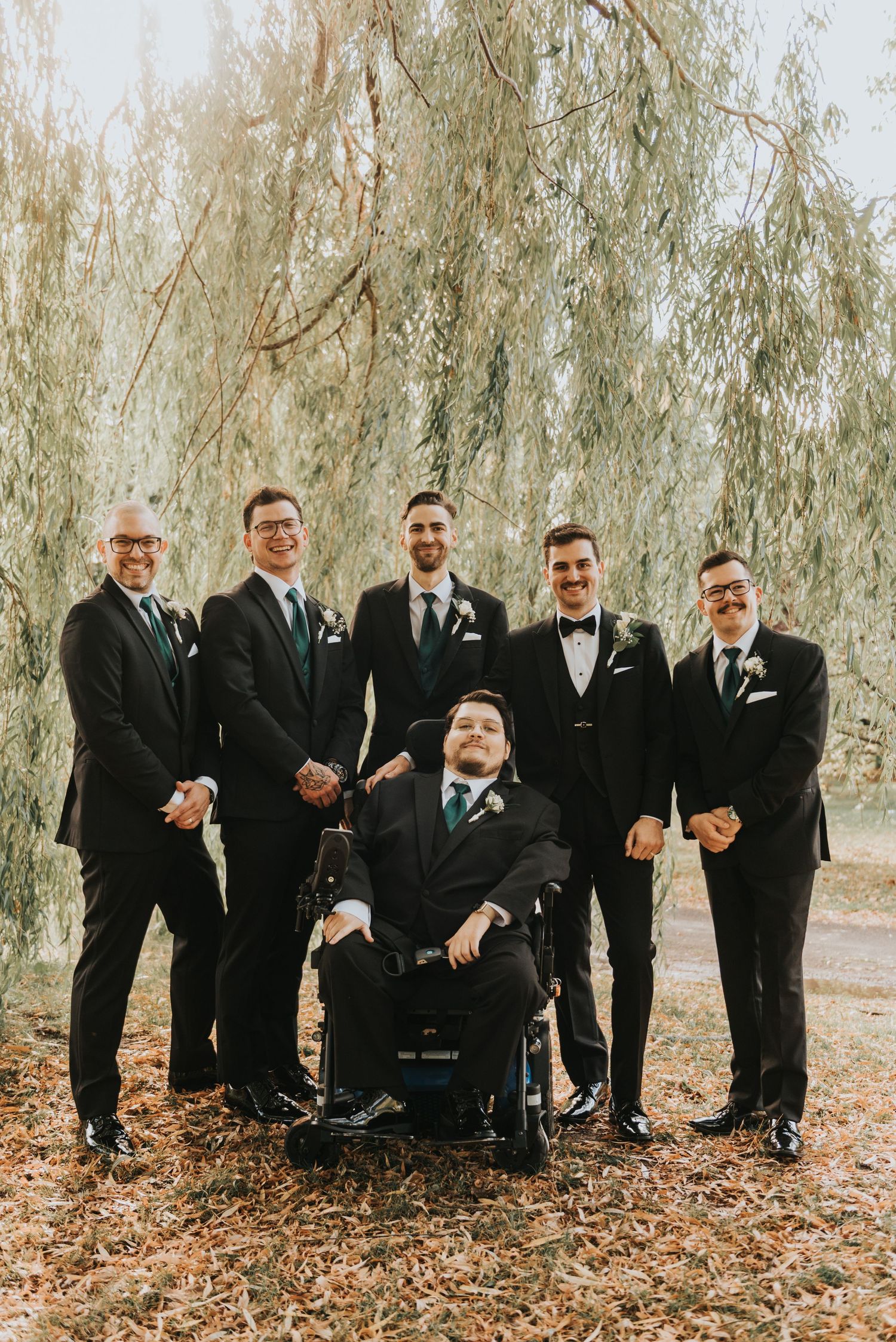 A group in black suits and bow ties poses together outdoors among willow trees on a fall day.