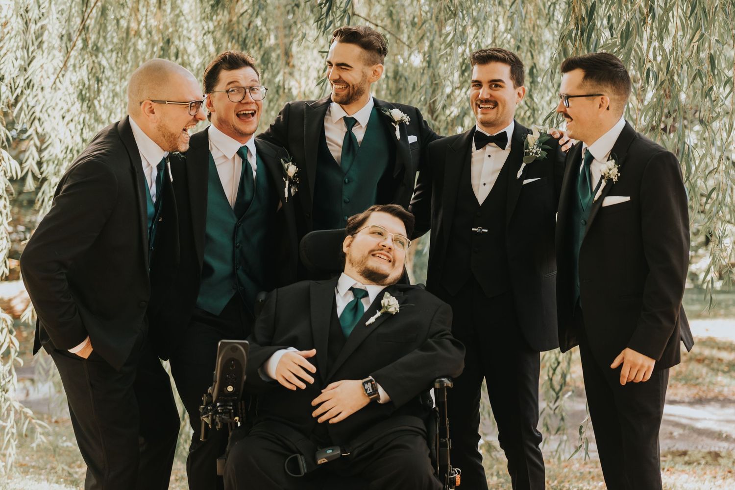 A wedding party laughs together while standing beneath drooping willow tree branches in black and white.