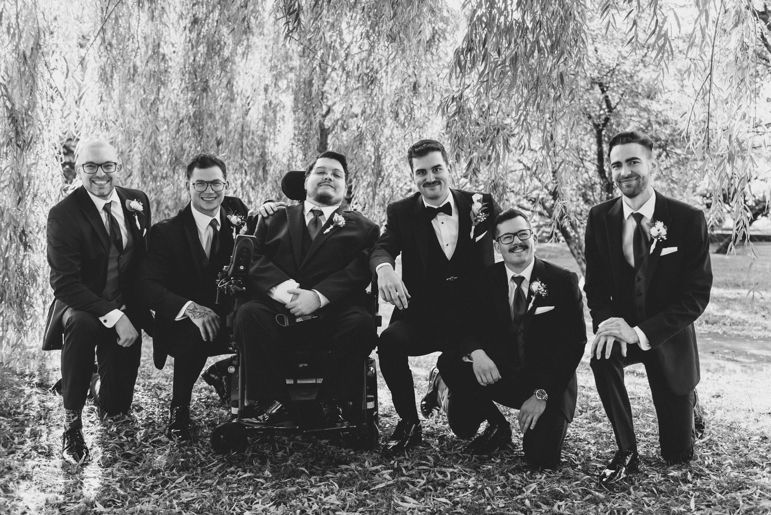 Black and white wedding party members sit together on the ground laughing joyfully in an outdoor setting.