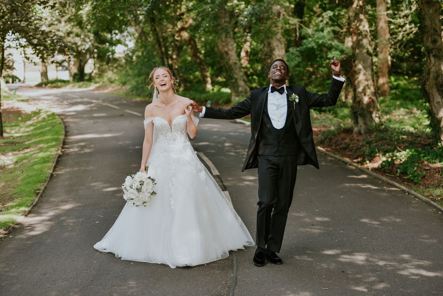 A couple in formal wedding attire walks along a tree-lined path during an outdoor ceremony with confetti at Brig o' doon Ayr