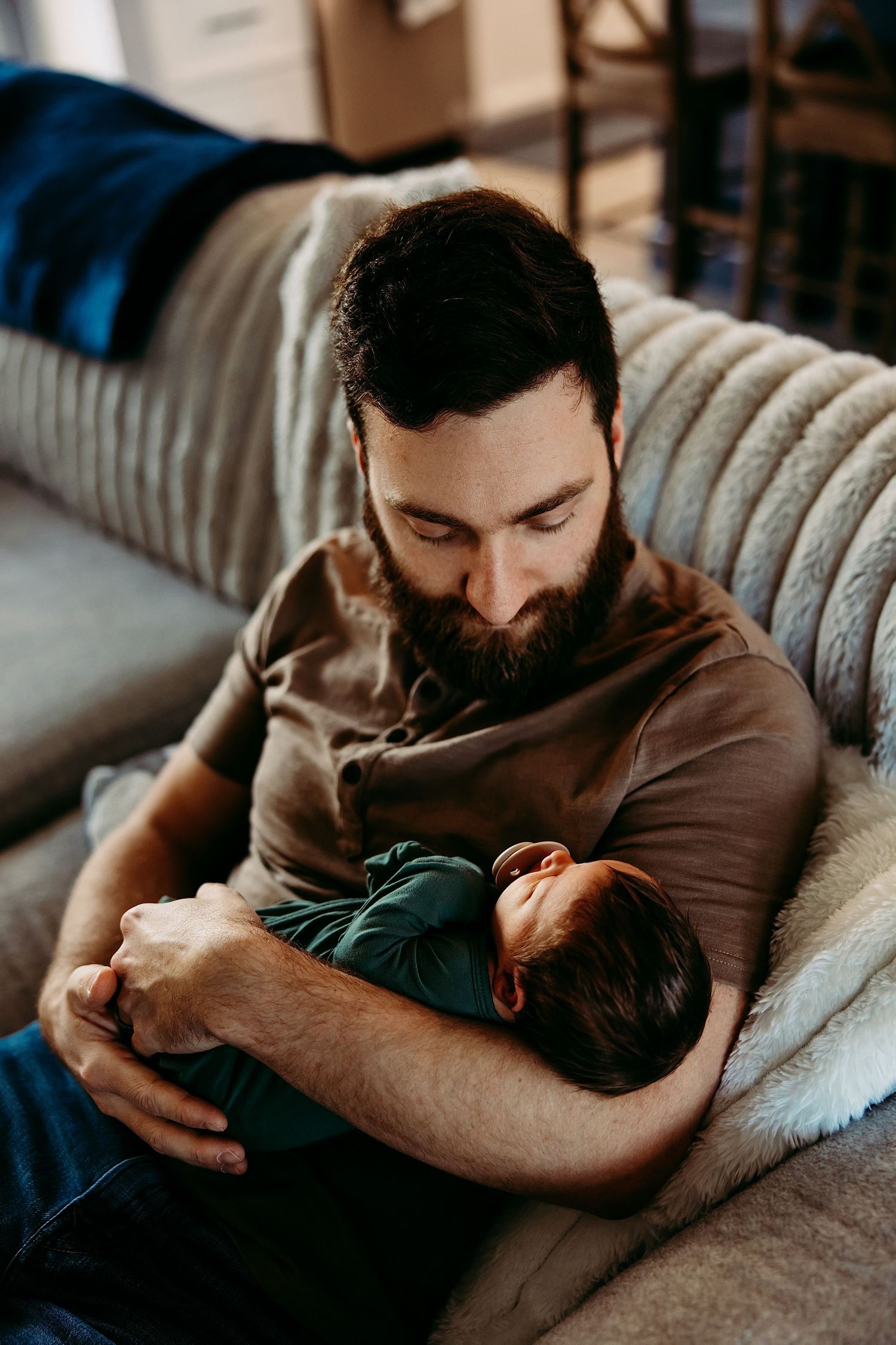 A bearded man in a brown shirt rests on a gray couch while holding a sleeping child in his arms.
