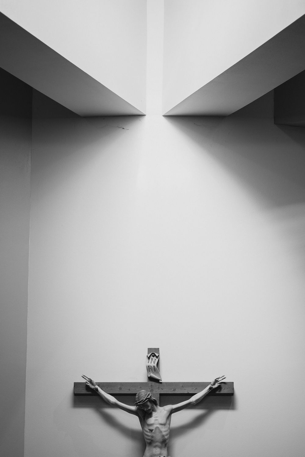 Black and white photo of a Jesus crucifix mounted on a white wall with dramatic angular ceiling light.