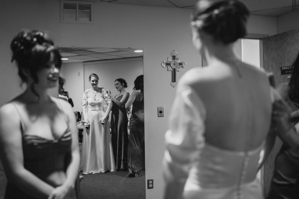 Bride in white gown views reflection in mirror surrounded by bridesmaids in black and white wedding preparation photo.