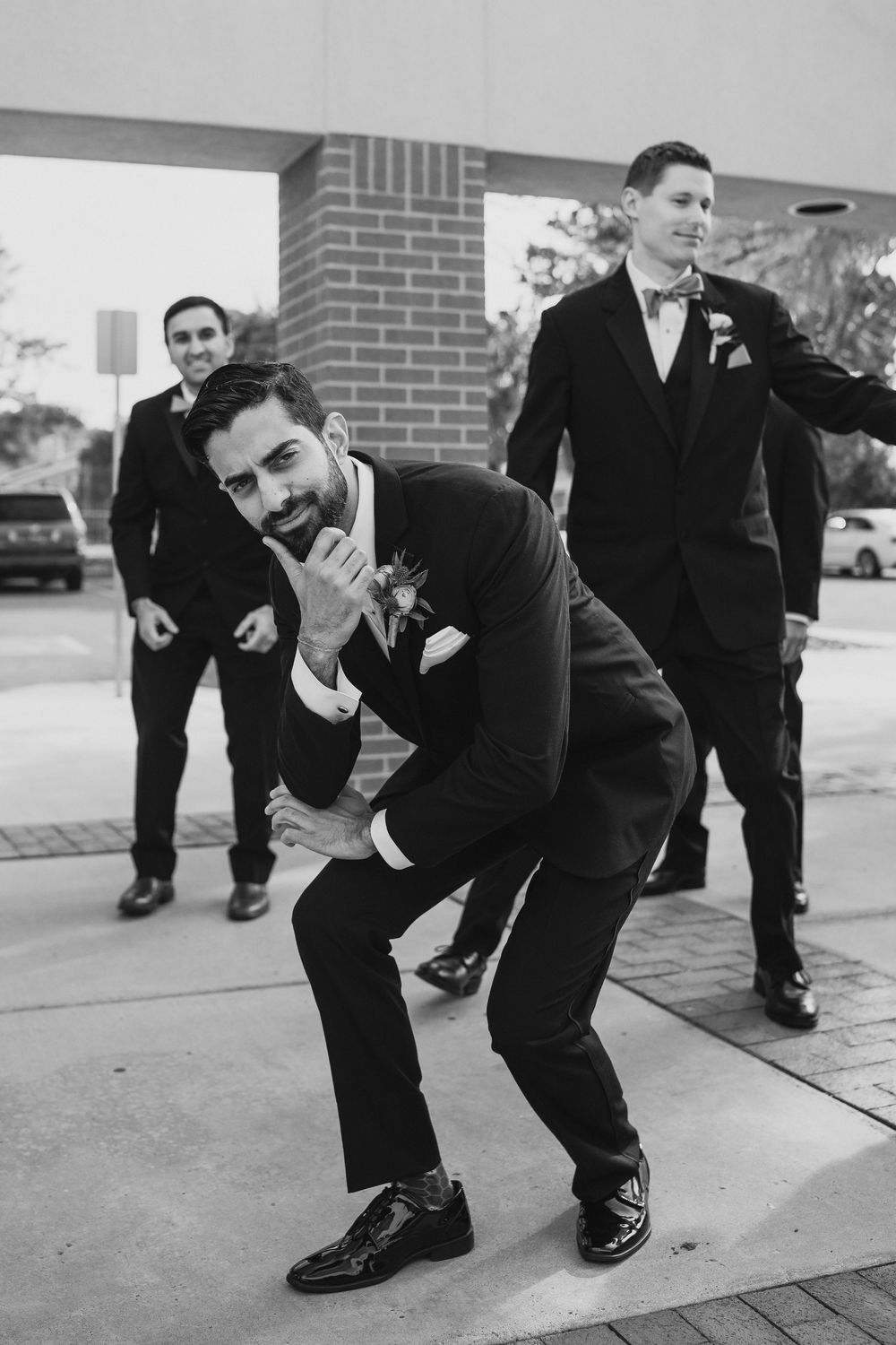 Groom and groomsmen in black tuxedos strike fun poses outside brick building in black and white wedding photo.