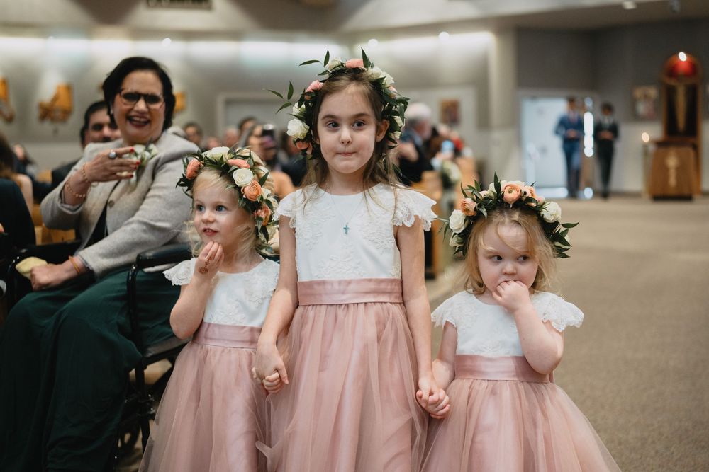 Three flower girls in pink dresses and floral crowns stand together during wedding ceremony in church.