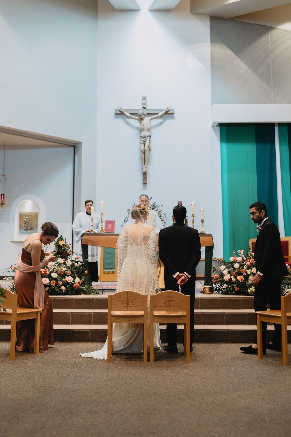 Bride and groom kneel at altar before crucifix during Catholic wedding ceremony with wedding party.