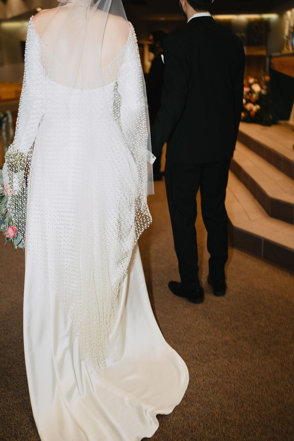 Close-up back view of bride in beaded long-sleeve wedding gown with veil walking toward altar.