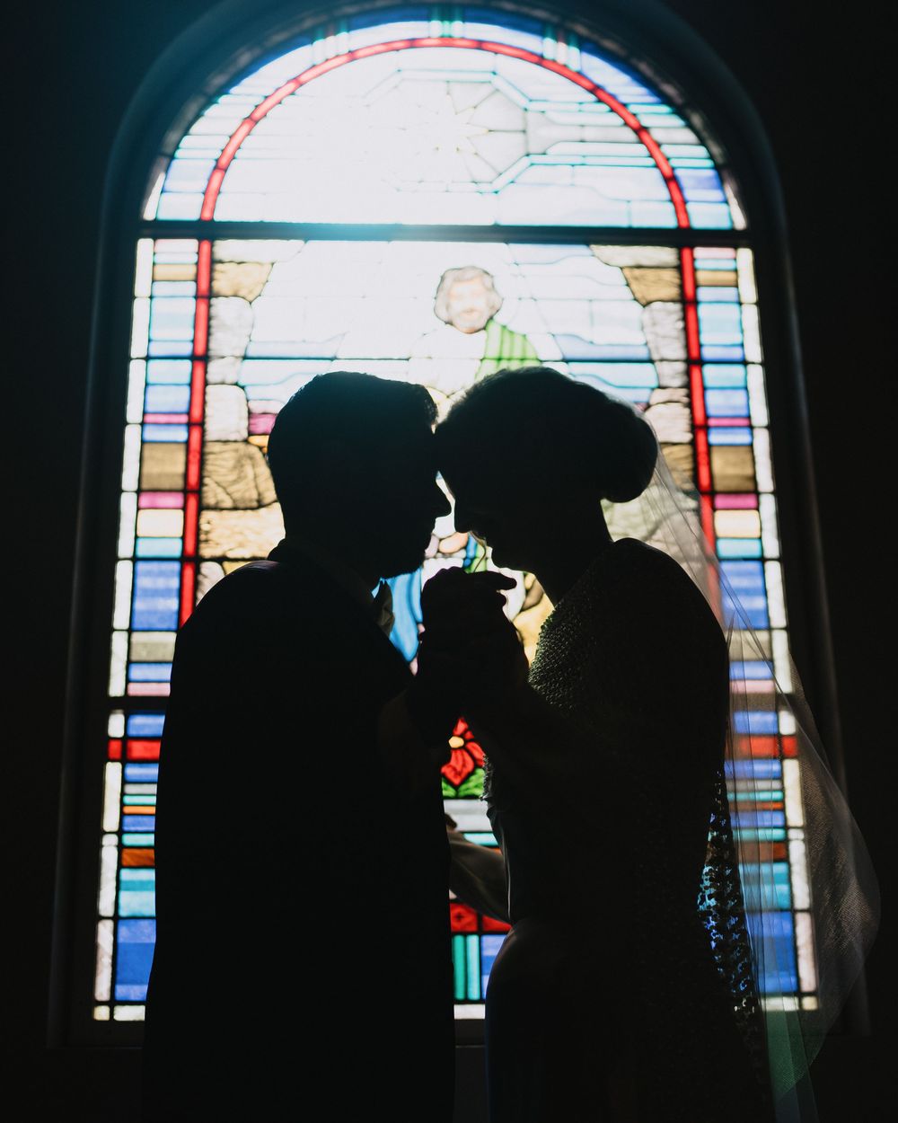 Romantic silhouette of bride and groom kissing in front of colorful stained glass church window.