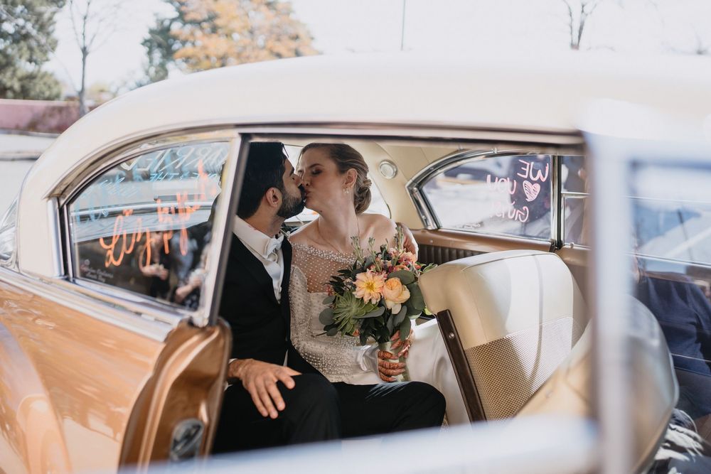 Bride and groom share a kiss inside vintage classic car after wedding ceremony with bouquet.