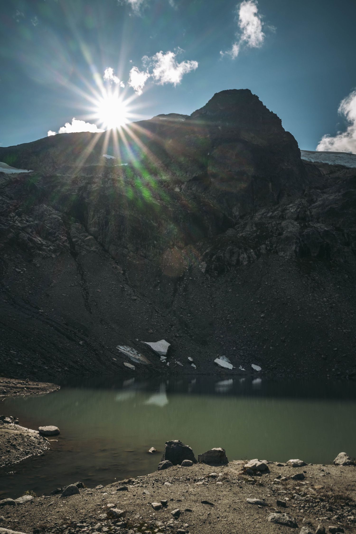 Sun flare over rocky mountain peak reflecting in alpine lake with patches of snow.