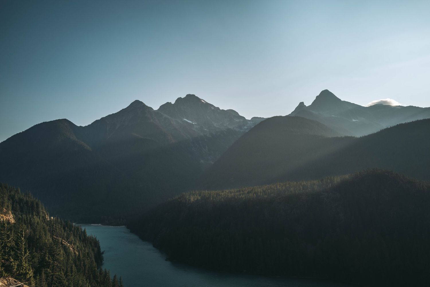 Panoramic view of layered mountain peaks above forested valley with calm lake below.