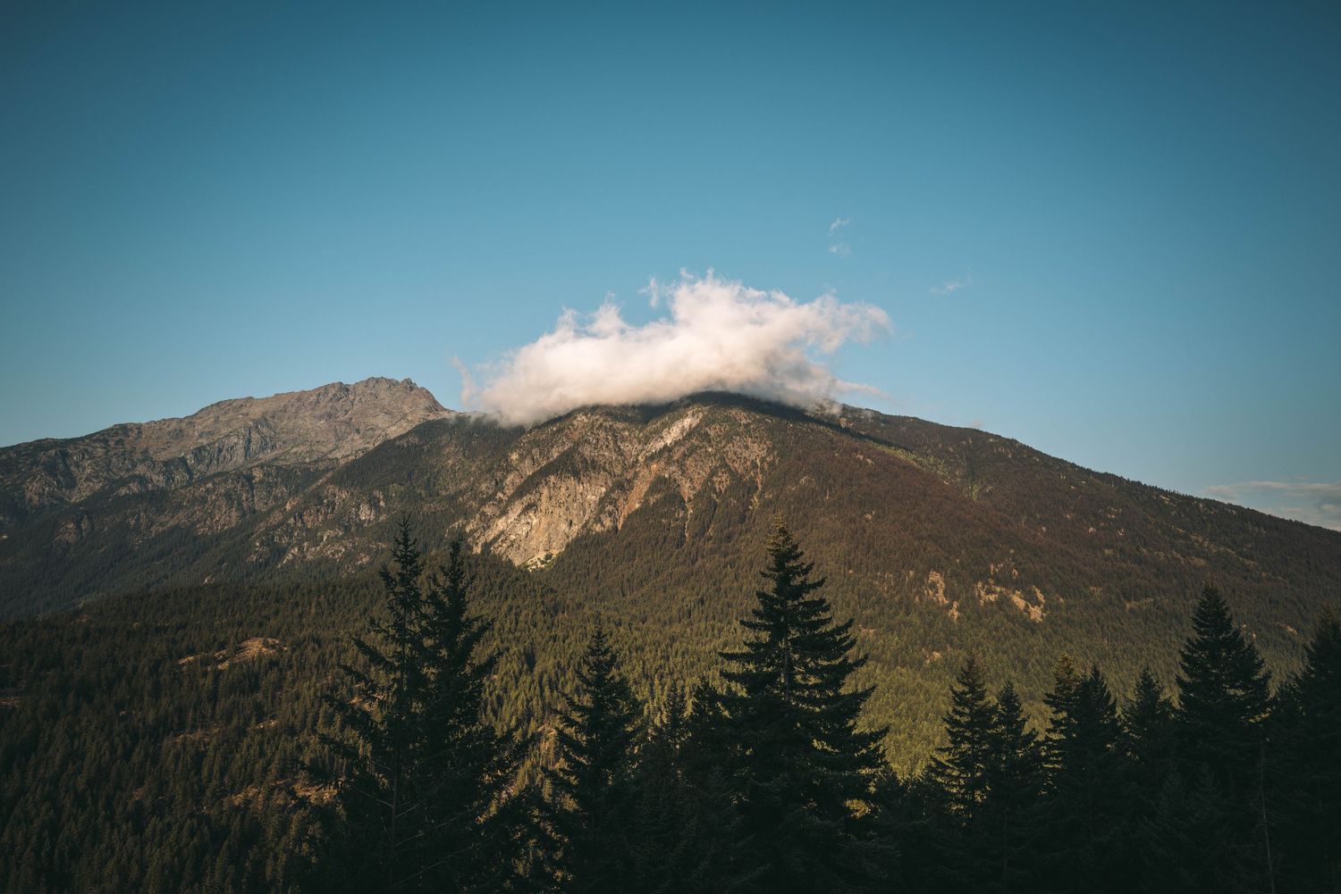 Mountain summit with dramatic cloud formation above pine forest under clear blue sky.