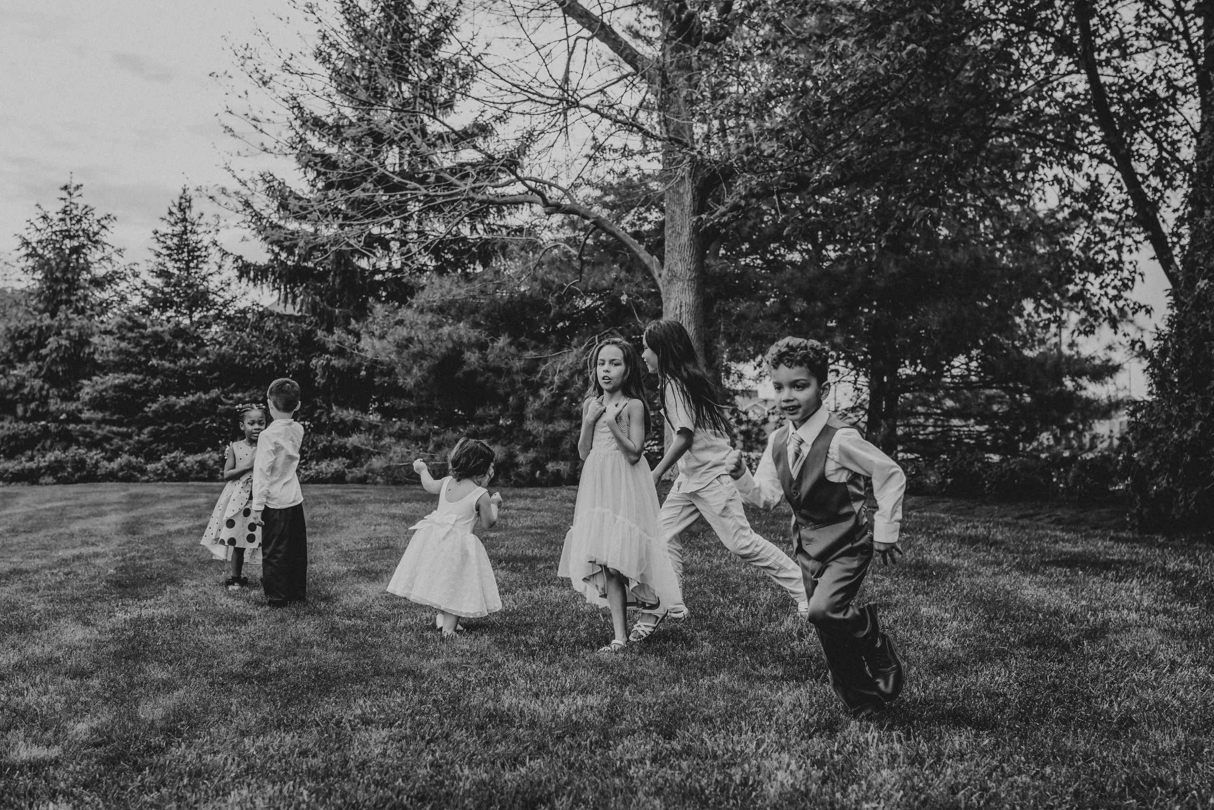 Black and white photo of people dancing and celebrating on a grassy lawn with evergreen trees in the background.