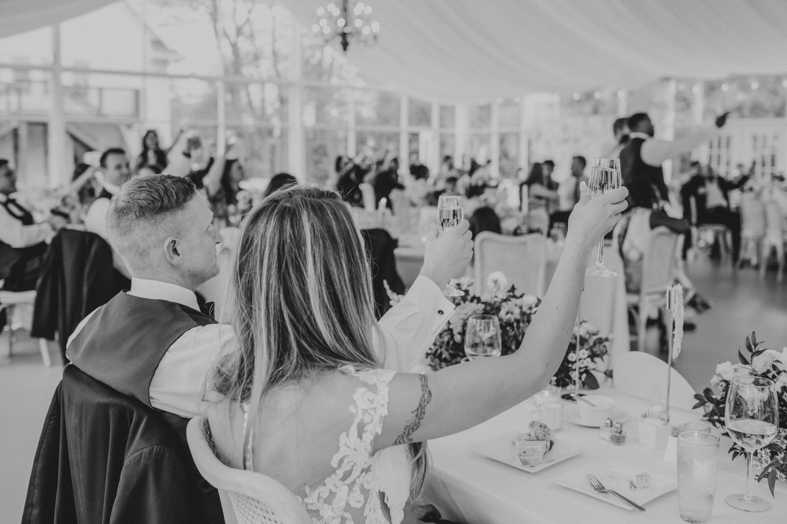 Wedding reception guests raise their glasses in a toast at an elegant table setting under a white tent.