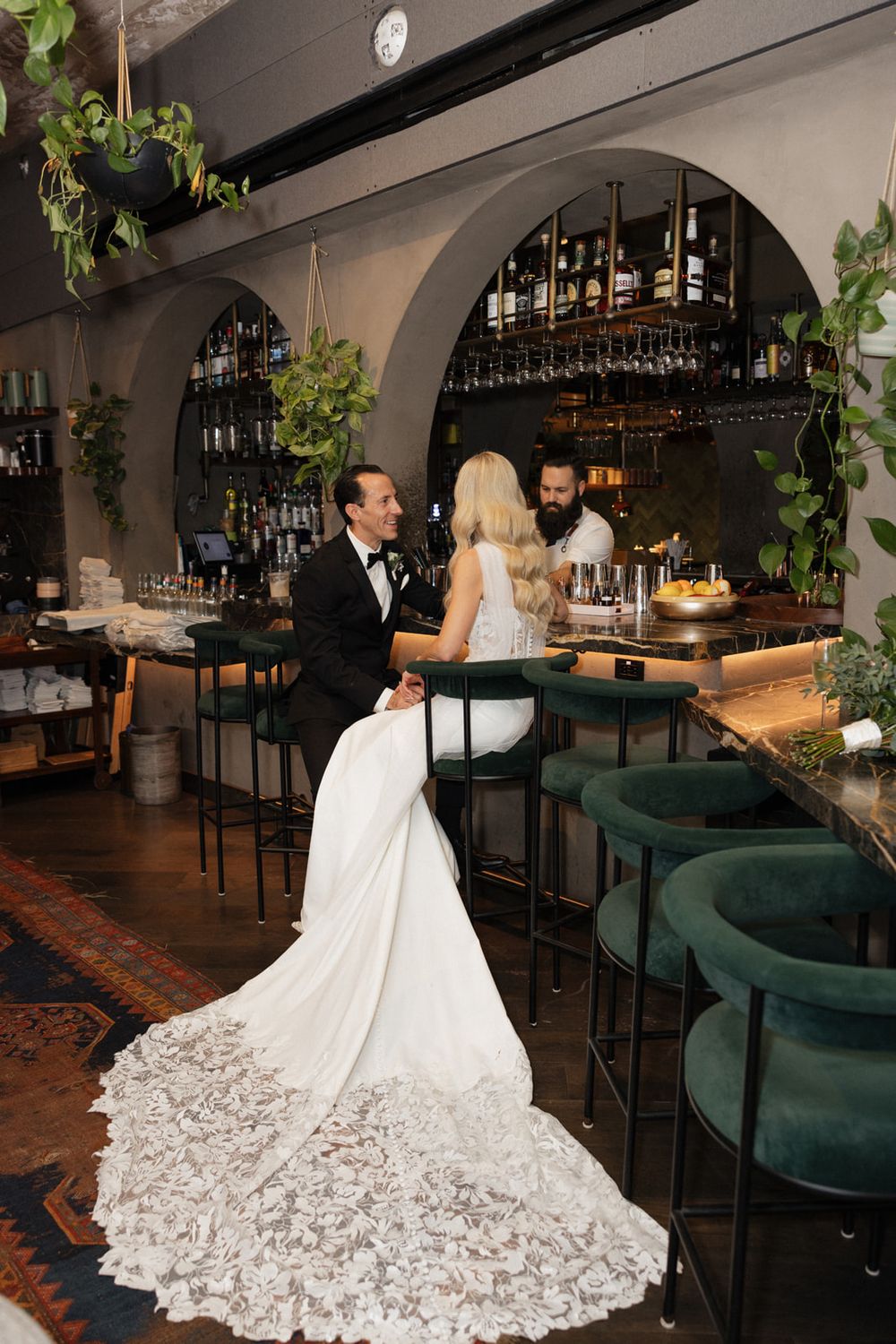 Elegant wedding dress with intricate lace train sweeps across a restaurant floor with emerald green seating and bar decor.