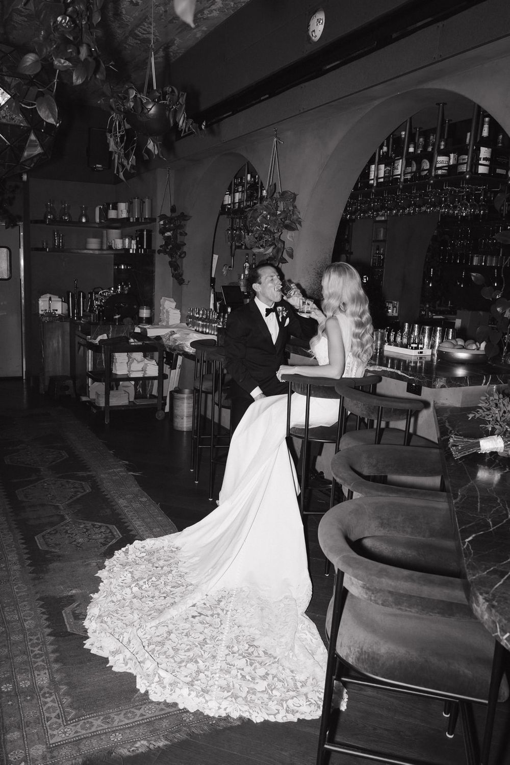 A couple in formal wedding attire share an intimate moment at an elegant vintage bar with a dramatic lace train.