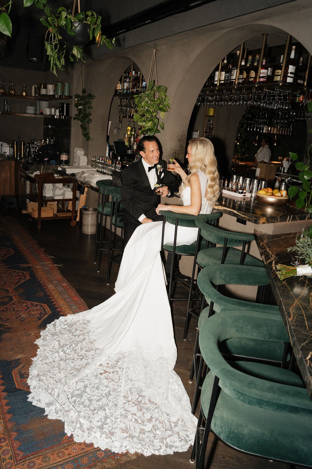 A couple in wedding attire sits at an elegant bar with green velvet stools and a dramatic lace train on the white gown.
