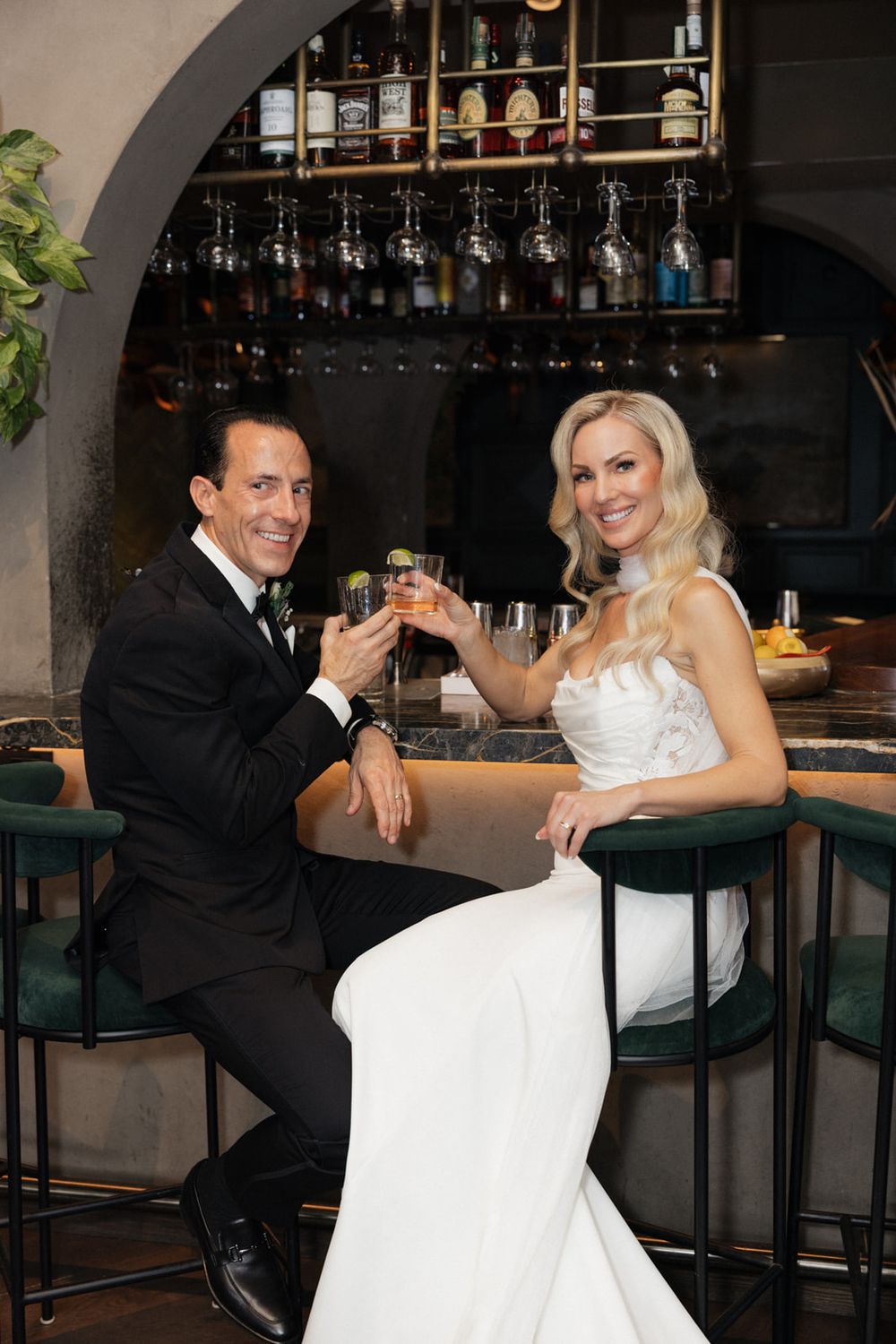 A couple in wedding attire shares playful moments at a bar counter with liquor bottles displayed in the background.
