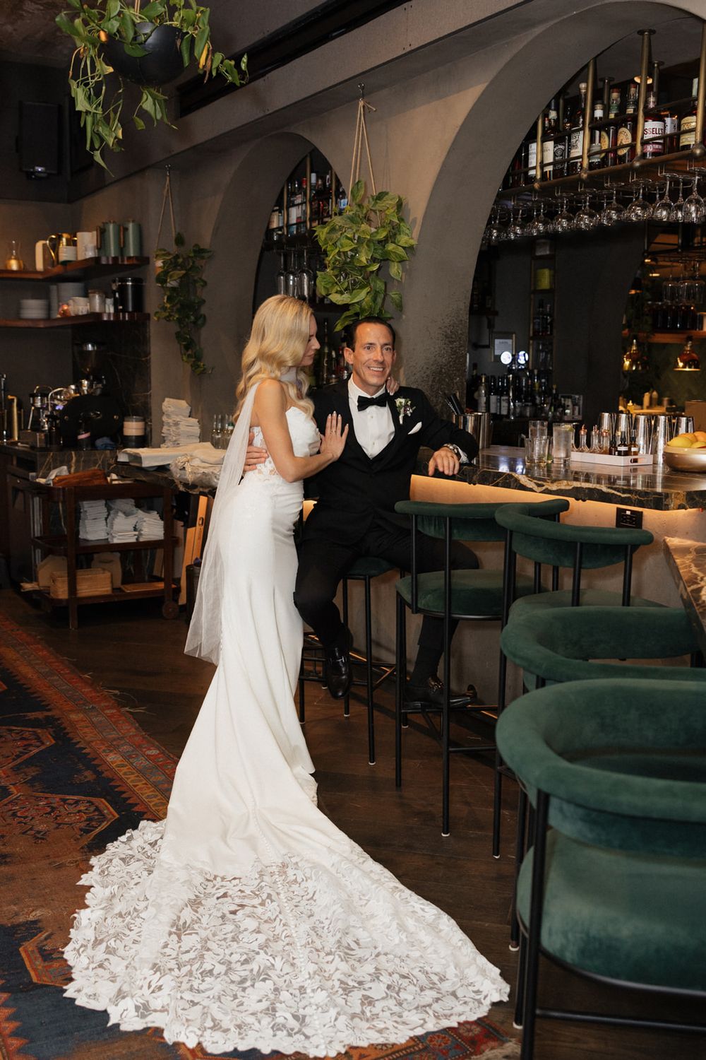 A bride in a flowing white lace gown with a dramatic train poses in an elegant restaurant setting with emerald velvet seating.
