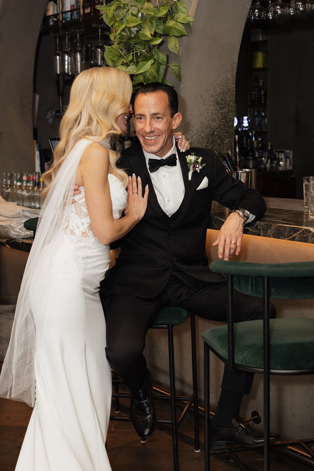 Bride in white dress and groom in tuxedo share intimate moments at a stylish bar during their wedding celebration.